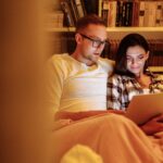 A couple enjoying a cozy evening reading together on a tablet in a home library, symbolizing quiet connection and understanding different social needs in relationships.