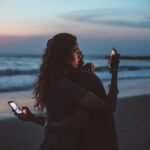 Couple sitting apart on seashore at sunset, both distracted by their smartphones instead of connecting with each other