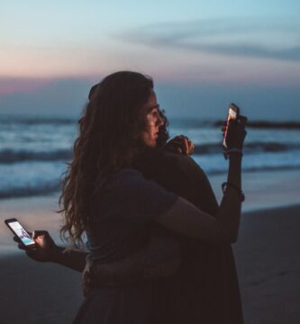 Couple sitting apart on seashore at sunset, both distracted by their smartphones instead of connecting with each other