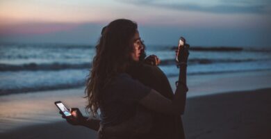Couple sitting apart on seashore at sunset, both distracted by their smartphones instead of connecting with each other