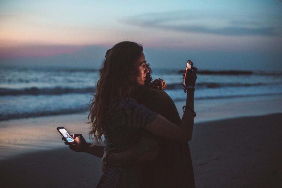 Couple sitting apart on seashore at sunset, both distracted by their smartphones instead of connecting with each other