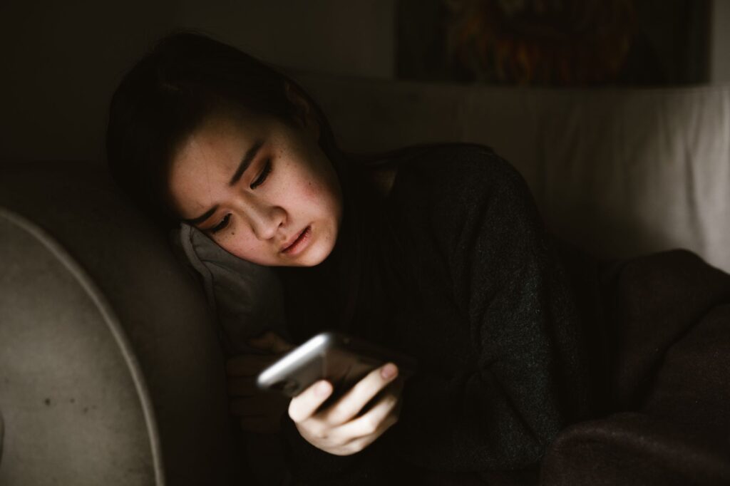 A young woman lying on a couch looking at her smartphone in a dimly lit room, illustrating the experience of retroactive jealousy and obsessive thoughts.