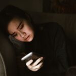 A young woman lying on a couch looking at her smartphone in a dimly lit room, illustrating the experience of retroactive jealousy and obsessive thoughts.