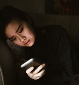 A young woman lying on a couch looking at her smartphone in a dimly lit room, illustrating the experience of retroactive jealousy and obsessive thoughts.