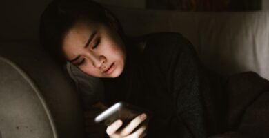 A young woman lying on a couch looking at her smartphone in a dimly lit room, illustrating the experience of retroactive jealousy and obsessive thoughts.